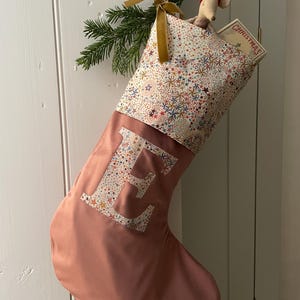 May include: A Christmas stocking in dusty rose fabric featuring a large letter E appliqued in a multi-coloured star pattern. The cuff displays the same star print with a gold velvet ribbon bow, a small mouse figurine, and a book labelled CHRISTMAS.