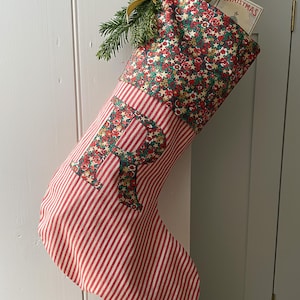 May include: A fabric Christmas stocking featuring a red and white vertical striped body with a floral patterned cuff. The letter R is appliqued on the front in the same floral fabric. A gold ribbon and pine sprig are attached to the top corner.