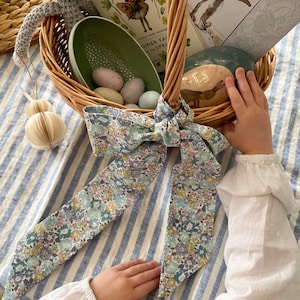 May include: A wicker Easter basket filled with books, decorative eggs, and a crocheted bunny. A floral patterned ribbon is tied to the basket handle. The basket is on a blue and white striped surface.