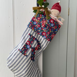 Op de afbeelding: Een stoffen kerstkous met een blauw-wit gestreept patroon en een grote letter R in bloemenprint. De boord is van bijpassende bloemenstof. Een kleine pop met een rode muts en een takje hulst met een gouden lint zijn in de kous gestoken.