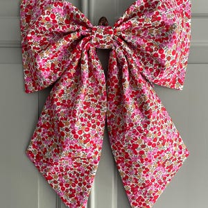 May include: A large fabric bow with a floral pattern in red, pink, and white. The bow is attached to a grey surface and has long, flowing tails. The floral pattern is made up of small, round shapes.
