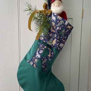 May include: A green fabric Christmas stocking featuring a large letter R appliqued in a dark blue fabric with a multi-coloured animal print. The cuff matches the letter fabric and holds a small fabric gnome doll, a pine sprig, and a gold ribbon.
