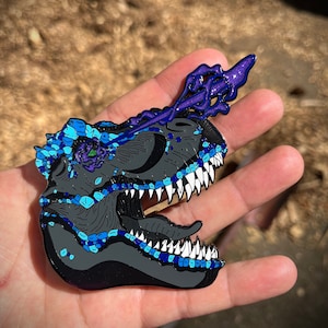 May include: A black enamel pin of a dinosaur skull with blue and purple glitter accents. The dinosaur's mouth is open, revealing sharp teeth. The pin is detailed with a floral design on the side of the skull.