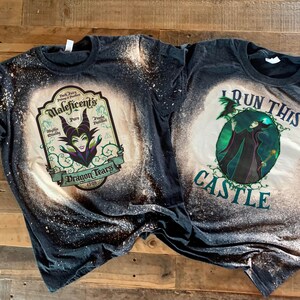 Op de afbeelding: Twee zwarte T-shirts met bleekspatten ontwerpen met Maleficent uit Disney's Doornroosje. E&eacute;n shirt zegt "I Run This Castle" en de andere zegt "Maleficent's Pure Dragon Tears".