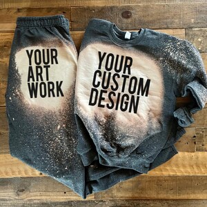 May include: A gray sweatshirt and sweatpants set with bleach splattered designs. The sweatshirt has the text "YOUR CUSTOM DESIGN" and the sweatpants have the text "YOUR ART WORK".