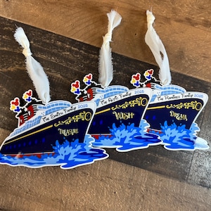 Puede incluir: Tres adornos en forma de barco azul marino con cintas blancas. Cada adorno tiene una palabra diferente: "Treasure", "Wish" y "Dream". Los adornos tienen el texto "The Smith Family 2025", "The Artic Family 2025" y "The Ramirez Family 2025".