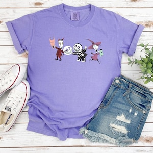 May include: Purple t-shirt with a graphic print of three characters from the Nightmare Before Christmas movie. The characters are Jack Skellington, Sally, and Oogie Boogie. The shirt is on a white wooden surface with a pair of white sneakers and denim shorts.