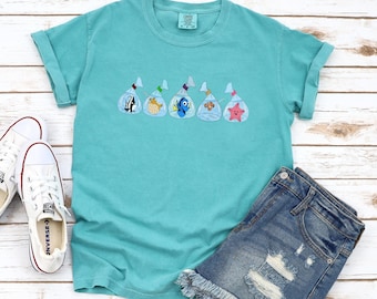 May include: A turquoise blue t-shirt with a graphic of five Finding Nemo characters in plastic bags. The characters are Dory, Marlin, Nemo, Squirt, and Pearl. The shirt is laid out on a white wood background with a pair of white sneakers and denim shorts.