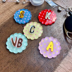 Puede incluir: Una colecci&oacute;n de coloridos carretes para insignias con forma de flor, con letras e iniciales. Cada carrete es de un color diferente, incluyendo azul, rojo, amarillo y morado, con lunares. Las letras son plateadas y doradas.