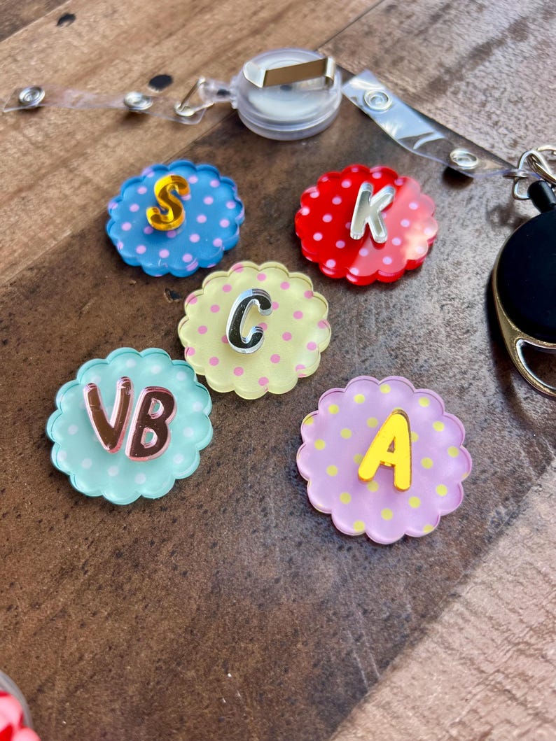 Puede incluir: Una colecci&oacute;n de coloridos carretes para insignias con forma de flor, con letras e iniciales. Cada carrete es de un color diferente, incluyendo azul, rojo, amarillo y morado, con lunares. Las letras son plateadas y doradas.