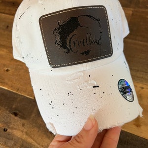May include: White baseball cap with black splatter paint and a brown leather patch with the word "Cruella" in black lettering.