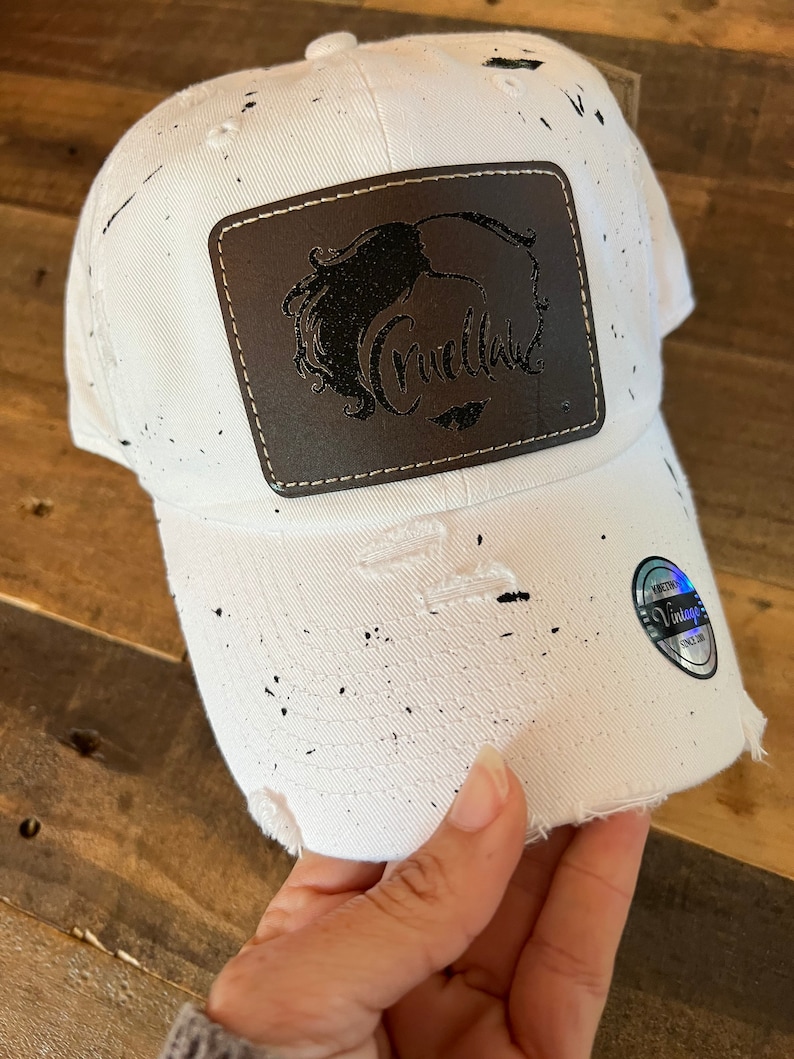 May include: White baseball cap with black splatter paint and a brown leather patch with the word "Cruella" in black lettering.