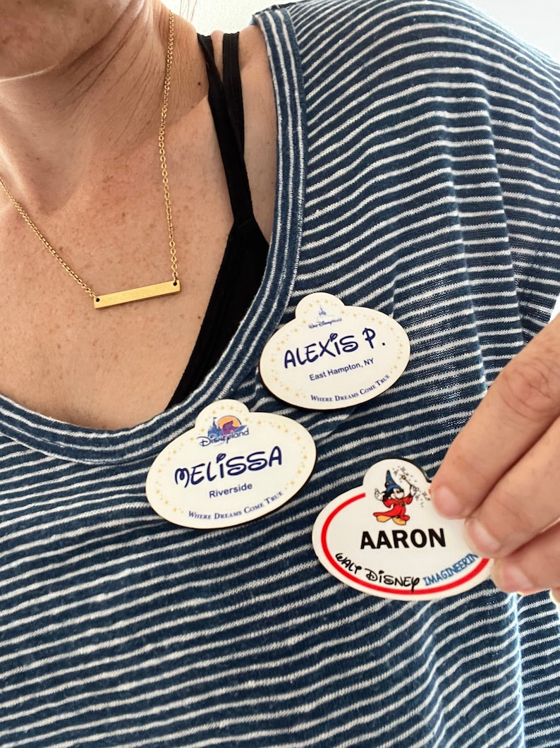 May include: Three Disney name tags with the names Alexis, Melissa, and Aaron. The name tags are oval-shaped with a white background and a red border. The image also shows a gold bar necklace and a blue and white striped shirt.
