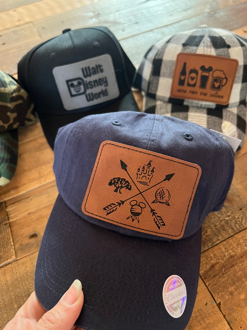 May include: Three baseball caps with different designs. The first cap is black with a white patch that says "Walt Disney World" and a Mickey Mouse silhouette. The second cap is white and black checkered with a brown patch that says "Here for the good times" and has a beer mug, a bottle, and a Mickey Mouse silhouette. The third cap is navy blue with a brown patch that has a Mickey Mouse silhouette, a castle, a tree, and a ball with a cross through it. The patch also has arrows pointing in different directions.