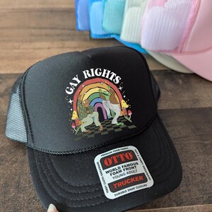 May include: Black trucker hat with the words "GAY RIGHTS" above a rainbow and cartoon figures. The hat has a mesh back and a snap closure. The hat is made of foam fabric.