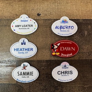 Peut inclure: Six badges Disneyland sont présentés. Les badges sont ovales avec des fonds blancs, affichant des noms, des lieux et des logos Disneyland. Un badge est rouge, avec un graphisme de Mickey Mouse et le texte "Guest of Honor".