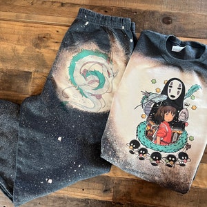 May include: A black and white bleached t-shirt and sweatpants set featuring a graphic print of characters from the animated film Spirited Away. The t-shirt features No Face, Chihiro, and the spirit of the river. The sweatpants feature a graphic print of the dragon Haku.