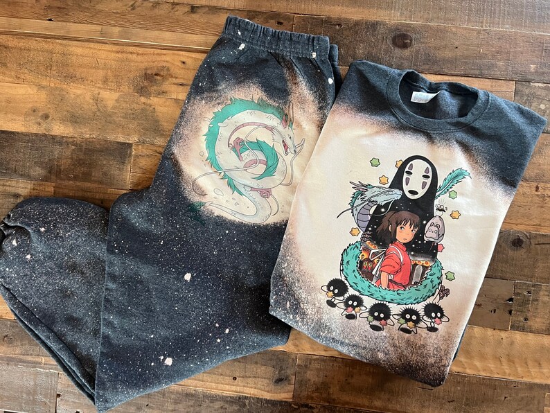 May include: A black and white bleached t-shirt and sweatpants set featuring a graphic print of characters from the animated film Spirited Away. The t-shirt features No Face, Chihiro, and the spirit of the river. The sweatpants feature a graphic print of the dragon Haku.