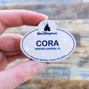 May include: A white and blue Walt Disney World name tag with the name "Cora" and location "Winter Garden, FL". The tag features a castle silhouette and the phrase "The Most Magical Place on Earth."