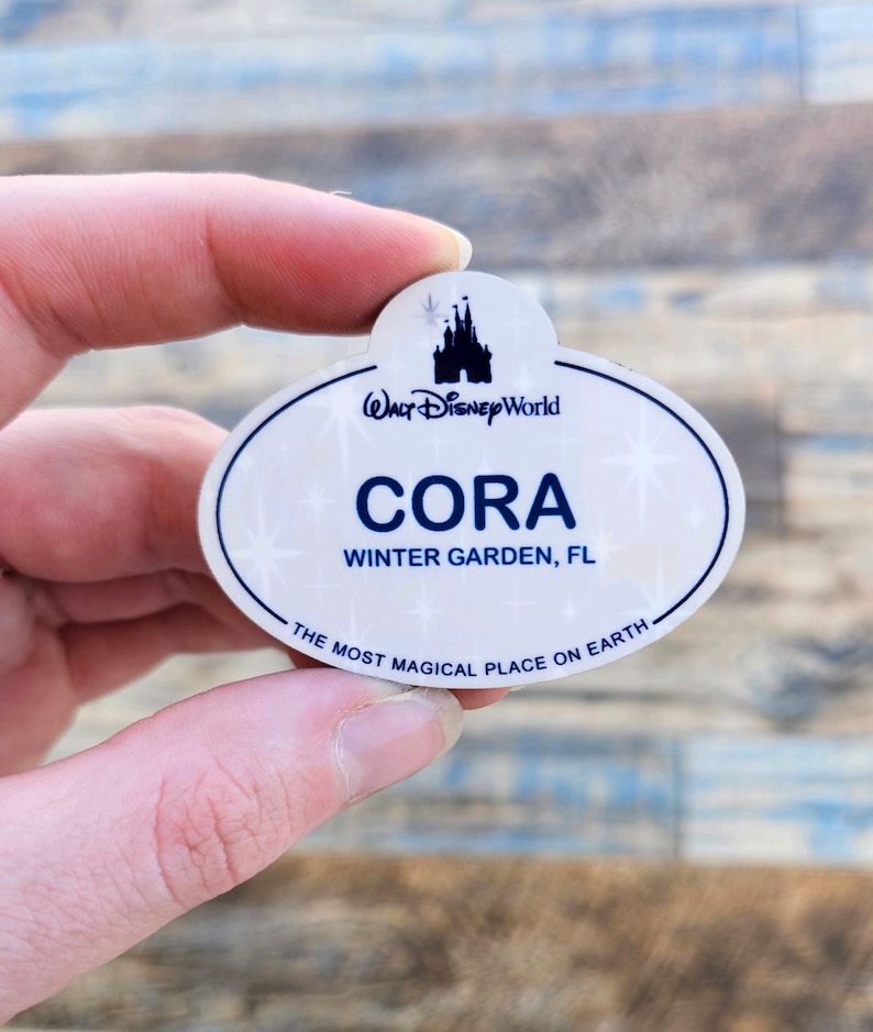 May include: A white and blue Walt Disney World name tag with the name "Cora" and location "Winter Garden, FL". The tag features a castle silhouette and the phrase "The Most Magical Place on Earth."