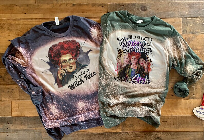Op de afbeelding: Twee shirts met lange mouwen met bleekmiddelspatten. Het eerste shirt is donkerblauw met een afbeelding van de Wicked Witch of the West uit de film The Wizard of Oz met de tekst "Resting Witch Face". Het tweede shirt is groen met een afbeelding van de drie heksen uit de film met de tekst "Oh look another Glorious Morning Makes Me Sick".