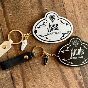 Könnte beinhalten: Zwei Schlüsselanhänger mit Namensschildern. Ein Schild ist weiß mit dem Namen "Jess" und den Worten "Ghost Host". Das andere Schild ist schwarz mit dem Namen "Nicole" und den Worten "Ghost Host". Jeder Schlüsselanhänger hat einen Sarganhänger.