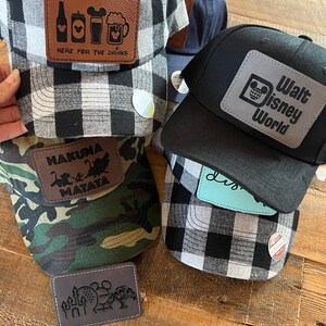 May include: Four different baseball caps with various patches. The first cap is black and white plaid with a brown leather patch that says "Here for the drinks" and has a beer mug, a can of beer, a bottle of beer, and a Mickey Mouse head. The second cap is black with a silver patch that says "Walt Disney World" and has a Mickey Mouse head. The third cap is camouflage with a brown leather patch that says "Hakuna Matata" and has a monkey and a lion. The fourth cap is black and white plaid with a brown leather patch that has a castle, a Mickey Mouse head, and a Ferris wheel.