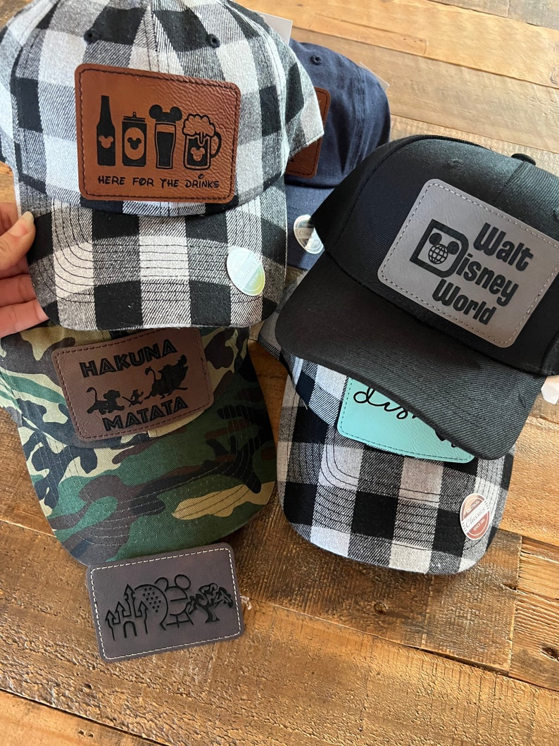 May include: Four different baseball caps with various patches. The first cap is black and white plaid with a brown leather patch that says "Here for the drinks" and has a beer mug, a can of beer, a bottle of beer, and a Mickey Mouse head. The second cap is black with a silver patch that says "Walt Disney World" and has a Mickey Mouse head. The third cap is camouflage with a brown leather patch that says "Hakuna Matata" and has a monkey and a lion. The fourth cap is black and white plaid with a brown leather patch that has a castle, a Mickey Mouse head, and a Ferris wheel.