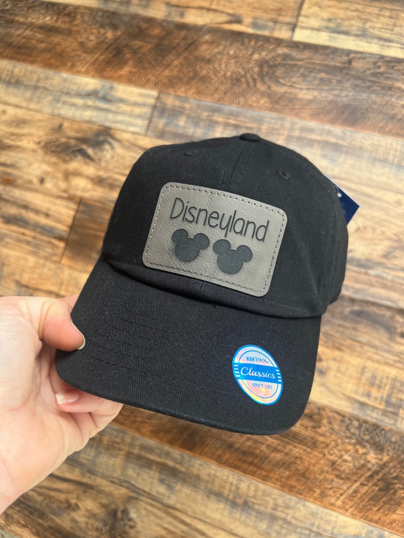 May include: Black baseball cap with a gray leather patch that says "Disneyland" with Mickey Mouse ears. The cap has a blue and white label that says "Kith & Kin Classics Since 1994."