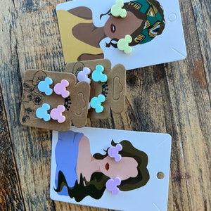 May include: A set of colorful Mickey Mouse-shaped earrings on a card with a woman's illustration. The earrings are in shades of pink, blue, and yellow.