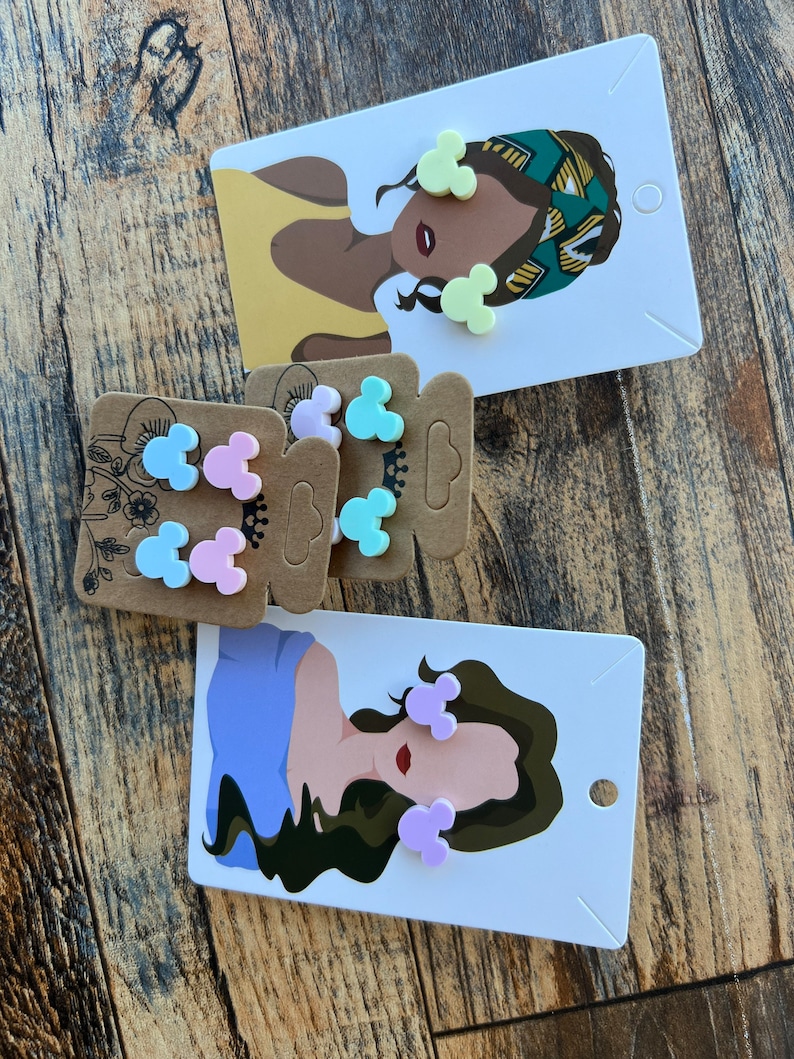 May include: A set of colorful Mickey Mouse-shaped earrings on a card with a woman's illustration. The earrings are in shades of pink, blue, and yellow.