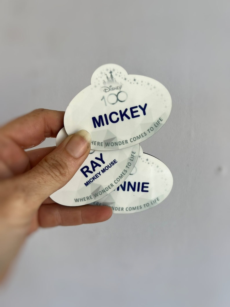 May include: Oval-shaped Disney name tags with the text "MICKEY", "RAY", and "NNIE" in navy blue. The tags have a white background with the Disney 100 logo and the phrase "WHERE WONDER COMES TO LIFE".