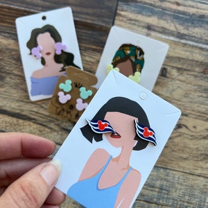 May include: A set of three pairs of earrings featuring a cartoon mouse head design. The earrings are displayed on white cards with illustrations of people wearing the earrings. The earrings are available in pink, blue, and yellow.