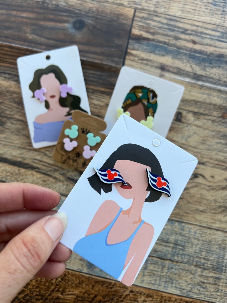 May include: A set of three pairs of earrings featuring a cartoon mouse head design. The earrings are displayed on white cards with illustrations of people wearing the earrings. The earrings are available in pink, blue, and yellow.