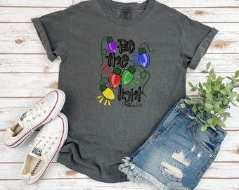 Peut inclure: Un t-shirt gris foncé avec un motif de lampes colorées et le texte "Soyez la lumière, Matthieu 5:14".
