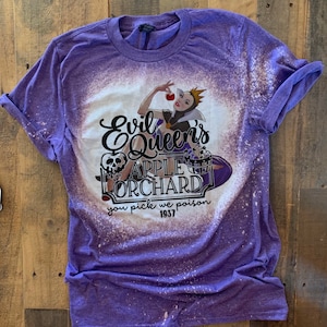 Op de afbeelding: Paars T-shirt met een ontwerp met bleekmiddelspatten met een illustratie van de boze koningin uit Sneeuwwitje die een rode appel vasthoudt. De tekst "Evil Queen's Apple Orchard you pick we poison 1937" is op het shirt gedrukt.