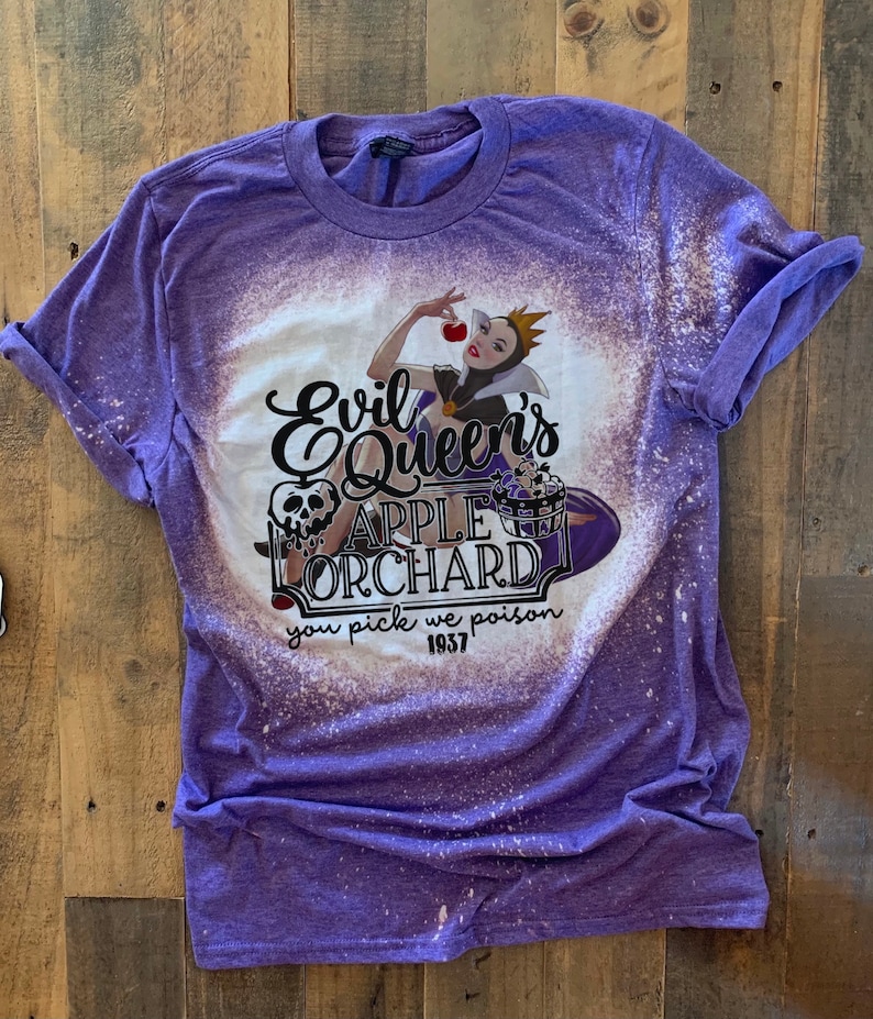 Op de afbeelding: Paars T-shirt met een ontwerp met bleekmiddelspatten met een illustratie van de boze koningin uit Sneeuwwitje die een rode appel vasthoudt. De tekst "Evil Queen's Apple Orchard you pick we poison 1937" is op het shirt gedrukt.