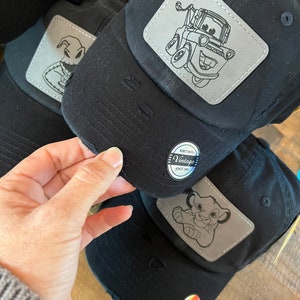 May include: Three black baseball caps with distressed fabric. Each cap features a different embroidered patch: a character from Nightmare Before Christmas, Mater from Cars, and Simba from The Lion King. Each patch is silver with black outlines. The caps have a black and white label that reads "Krethons Vintage Since 2001".