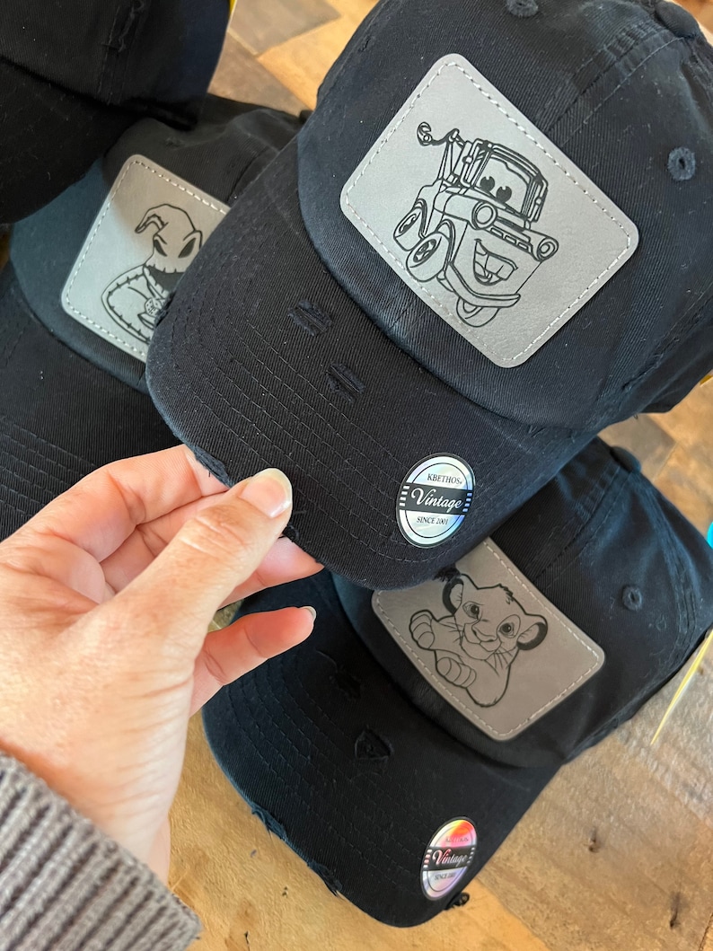 May include: Three black baseball caps with distressed fabric. Each cap features a different embroidered patch: a character from Nightmare Before Christmas, Mater from Cars, and Simba from The Lion King. Each patch is silver with black outlines. The caps have a black and white label that reads "Krethons Vintage Since 2001".
