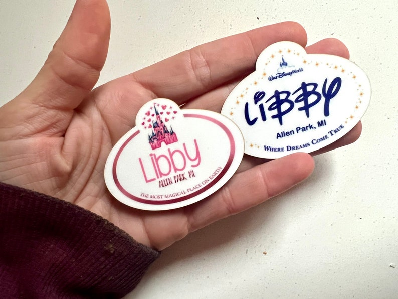 May include: Two oval-shaped name tags with the name "Libby". One tag features a pink castle graphic and the text "The Most Magical Place on Earth". The other tag has the text "Where Dreams Come True".