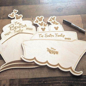 May include: Two wooden cruise ship-shaped signs with engraved text, including "The Benida Family 2025" and "The Smith Family 2025". The signs feature Mickey Mouse head cutouts and the words "Dream" and "Disney Treasure". A black marker is also present.