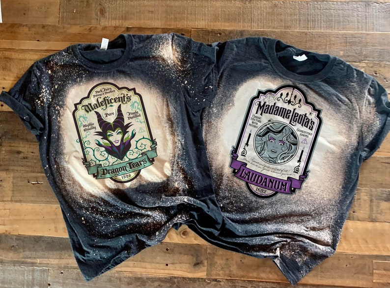 Op de afbeelding: Twee zwarte T-shirts met bleekspatten-ontwerpen. Het linker T-shirt heeft een vintage-stijl label met de tekst "Maleficent's Pure Dragon Tears 1698" en een illustratie van Maleficent. Het rechter T-shirt heeft een vintage-stijl label met de tekst "Madame Leota's Laudanum 1692" en een illustratie van Madame Leota.