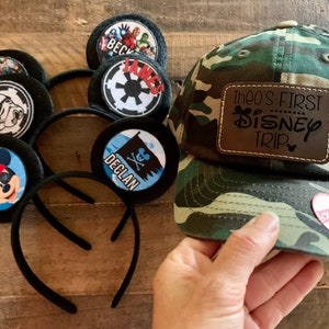 May include: A set of five black mouse ears with different character designs, including Mickey Mouse, Star Wars, and Avengers. There is also a camouflage baseball cap with a brown leather patch that says "Theo's First Disney Trip".