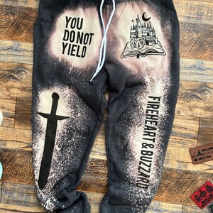 May include: Black sweatpants with a white bleach design. The design includes a castle, a book, a sword, and the text "You Do Not Yield" and "Fireheart & Buzzard".