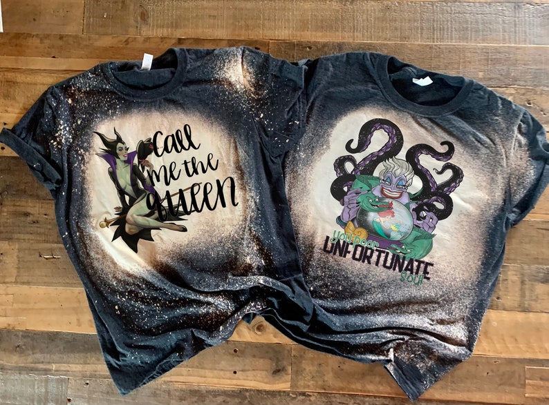 Op de afbeelding: Twee zwarte T-shirts met bleekdesigns met Disney-schurken. Het eerste shirt heeft een bleekdesign van Maleficent uit Doornroosje met de tekst "Call me the Queen". Het tweede shirt heeft een bleekdesign van Ursula uit De Kleine Zeemeermin met de tekst "You poor unfortunate soul".