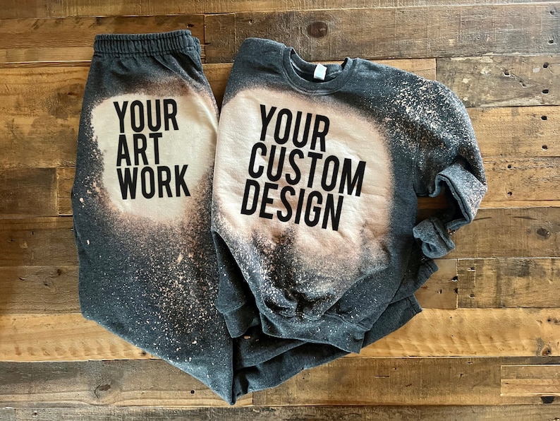 May include: A gray sweatshirt and sweatpants set with bleach splattered designs. The sweatshirt has the text "YOUR CUSTOM DESIGN" and the sweatpants have the text "YOUR ART WORK".
