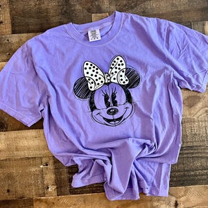 Puede incluir: Una camiseta morada con un dibujo en blanco y negro de Minnie Mouse con un lazo de lunares.