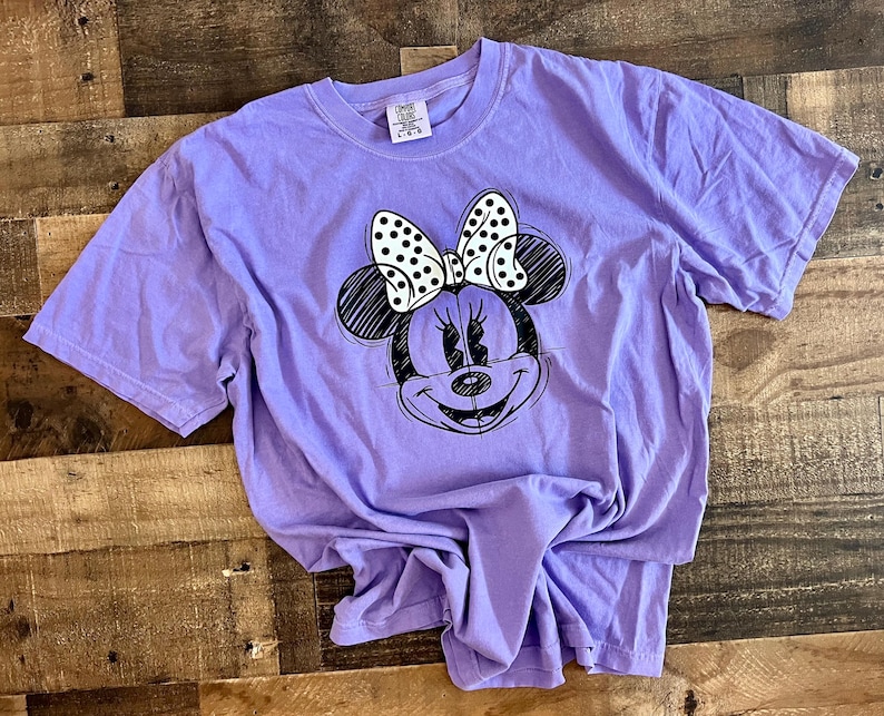 Puede incluir: Una camiseta morada con un dibujo en blanco y negro de Minnie Mouse con un lazo de lunares.