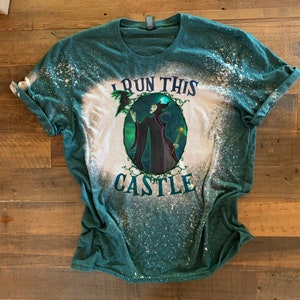 Op de afbeelding: Een groen T-shirt met een bleekmiddel-gespatte ontwerp met een cartoon afbeelding van Maleficent uit Disneys Doornroosje. De tekst "I run this castle" is op het shirt gedrukt.