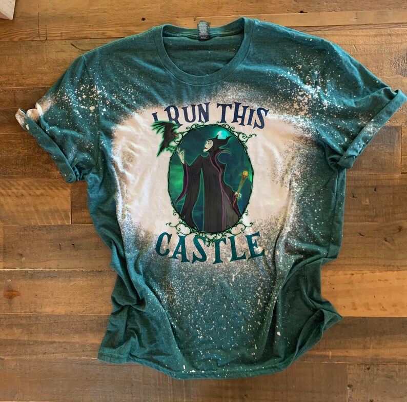 Op de afbeelding: Een groen T-shirt met een bleekmiddel-gespatte ontwerp met een cartoon afbeelding van Maleficent uit Disneys Doornroosje. De tekst "I run this castle" is op het shirt gedrukt.